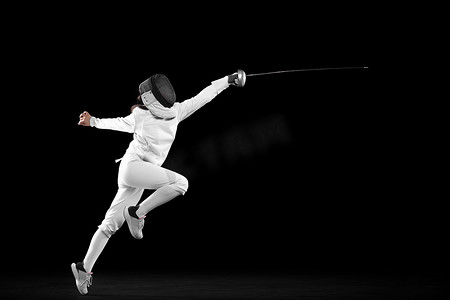 Energetic female fencer in white fencing costume and mask in action, motion isolated on dark background. Sport, youth, activity, skills, achievements. Girl practicing with rapier