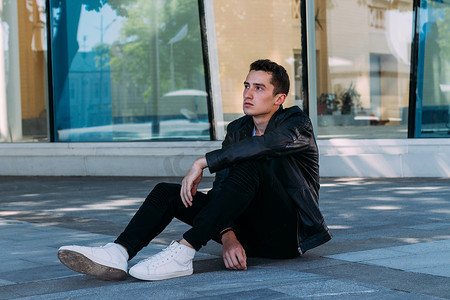 美丽和迷人的家伙坐在附近的玻璃建筑在城市的地板上, 摆姿势。身着黑色皮革衣服, 白色 t恤, 牛仔裤和白色鞋。城市风格