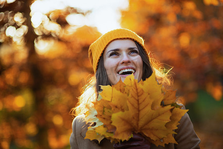 Hello autumn. smiling middle aged woman in beige coat and orange hat with autumn yellow leaves outside in the city park in autumn.