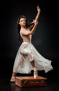 美丽的芭蕾舞演员在白色裙子站立与一条腿在复古手提箱在黑暗的背景上