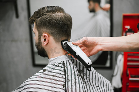 Professional male hairdresser does hairstyle to bearded man in barber shop, back view, barber's hand with clipper. Barber clipping client in men's hairdresser, closeup photo