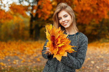 快乐的女人带着微笑在她的外衣里捧着一束秋天的黄叶在秋天公园的背景下