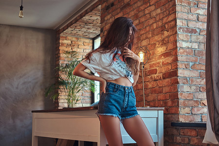性感苗条凉爽的女孩在白色的顶部和牛仔短裤, 站在一张桌子附近的阁楼内部的工作室.