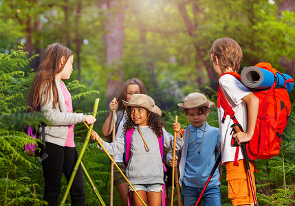 森林里走路摄影照片_一群小孩子在徒步旅行的路上互相交谈在森林里用杆子和背包走路
