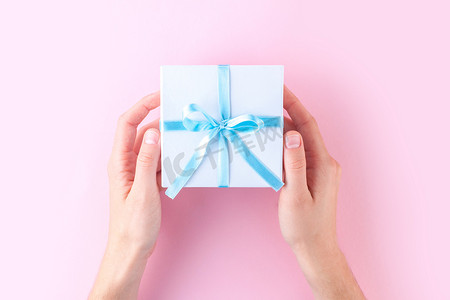 女性手拿着一个白色的小礼品盒包裹着蓝色的丝带在粉红色的背景。给予和接受来自亲人的礼物. 