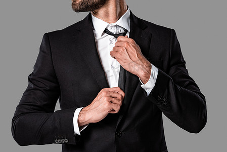 裁剪视图时尚英俊的商人在黑色西装触摸领带隔离在灰色