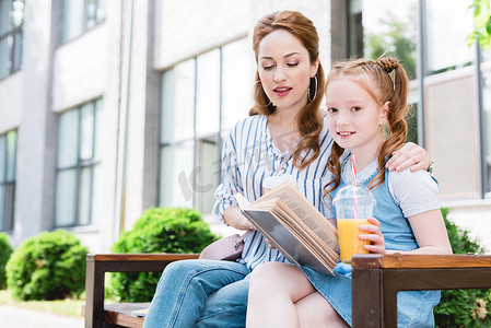 孩子的果汁阅读书与母亲附近, 而坐在长凳上一起在街上
