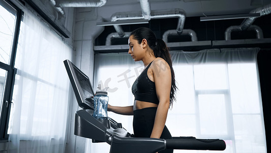 汗流浃背摄影照片_汗流浃背的女运动员在水瓶附近的跑步机上使用触摸屏