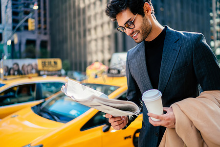 空白西装摄影照片_微笑成功的企业家在典雅的西装满意与文章在日报站立在大道与空白咖啡杯, 愉快的商人读报纸等待黄色出租汽车在 Manhatta