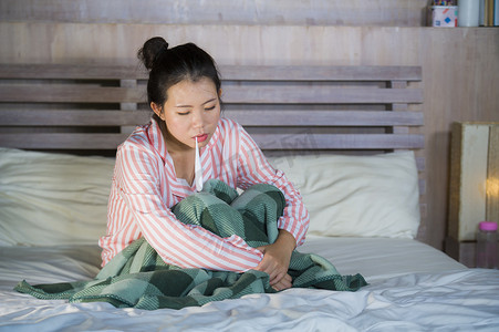 年轻美丽甜美的亚洲中国女孩穿着睡衣感到生病在家中卧室遭受感冒和流感的温度与温度计在床上覆盖着毯子与病毒感染