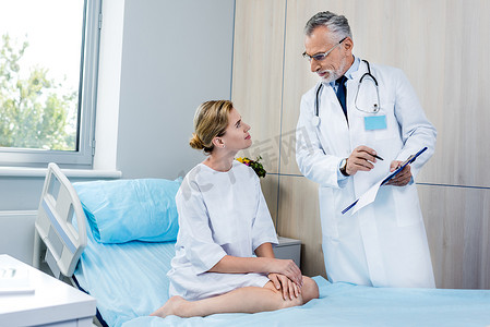 男性医生用听诊器在脖子指向在医院房间的女性患者的剪贴板 