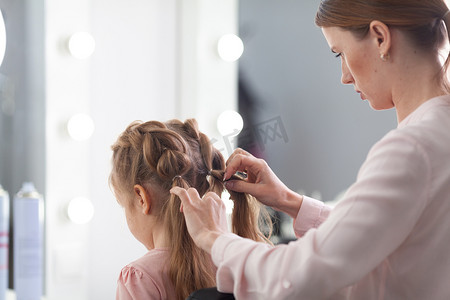 发型师在一个明亮的沙龙里做的发型设计，这个女孩在室内用一个漂亮的小模特做了卷发和辫子