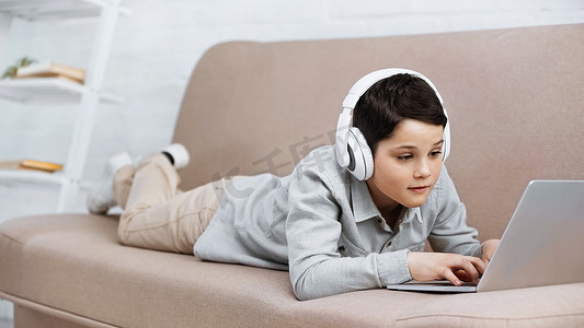 在沙发上使用笔记本电脑的戴耳机的未成年男孩  