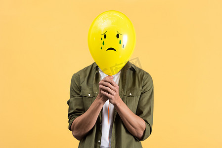 young man holding crying balloon in front of face, isolated on yellow