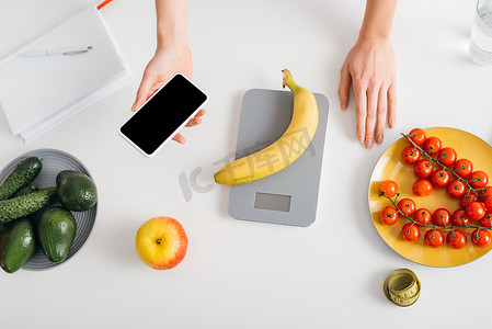 diet摄影照片_Top view of girl holding smartphone while weighing banana on kitchen table, calorie counting diet