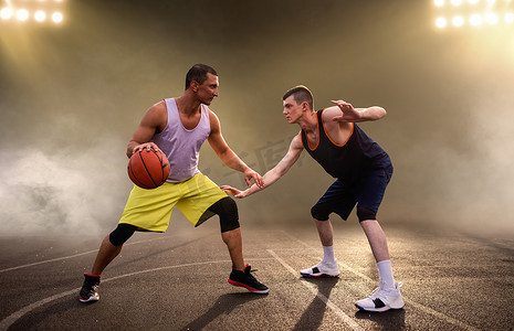 街头篮球摄影照片_两名篮球运动员在球场上打球，背景漆黑，灯光明亮。 男子运动员穿着运动服在街头篮球训练中比赛
