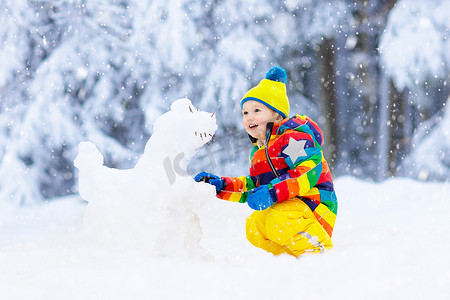 在雪冬公园里做雪人的孩子。孩子们在雪地里玩耍。小男孩在五颜六色的夹克和帽子大厦雪恐龙在冬天庭院在雪以后。冷冬日的户外娱乐.
