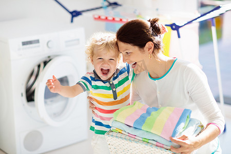 洗衣服摄影照片_母亲和孩子在洗衣房里用洗衣机或滚筒烘干机。家庭琐事。现代家庭设备和洗涤洗涤剂在白色阳光的家。烘干机架清洗洗衣服. 