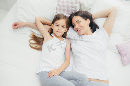 室内拍摄的欢快的母亲和女儿抱着躺在白色被褥上, 早上醒来, 有周末, 在卧室里, 有满意的表达。人, 生活方式概念