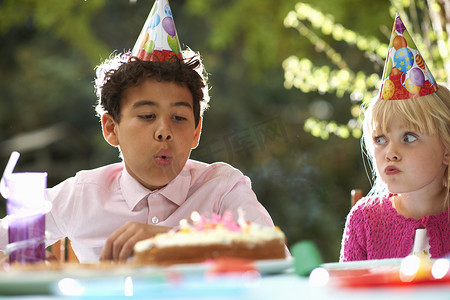 可爱生日蛋糕摄影照片_在花园生日聚会上男孩在生日蛋糕上吹蜡烛