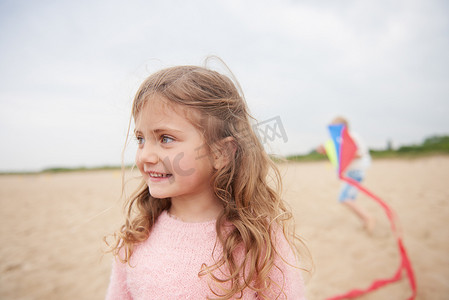 儿童动物背景摄影照片_小女孩在海滩上微笑背景中有放风筝的人