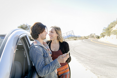 美国加利福尼亚州洛杉矶一对年轻夫妇靠在汽车上手持智能手机面对面微笑