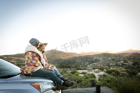 民族装饰边条摄影照片_坐在汽车引擎盖上面对面微笑的坐在汽车引擎盖上的山边年轻夫妇面带微笑美国洛杉矶加利福尼亚州奇劳露营地