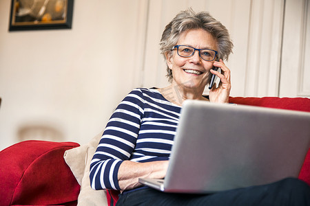 坐在客厅沙发上的资深女性一边使用笔记本电脑一边用智能手机交谈