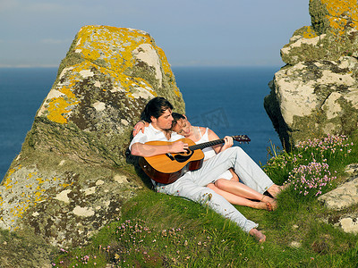 坐弹吉他摄影照片_年轻夫妇在悬崖上弹吉他