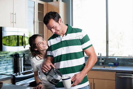 咖啡壶摄影照片_年轻夫妇在厨房里从壶里倒咖啡微笑着