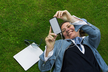 便携式摄影照片_俯瞰躺在草地上选择智能手机音乐的成熟女子