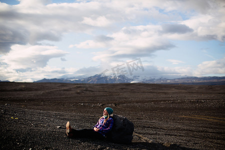 追逐摄影照片_中年妇女靠着巨石坐在火山景观上望向别处的侧视冰岛