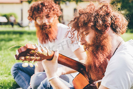 一对留着红胡子的年轻男性潮人双胞胎坐在公园里弹吉他