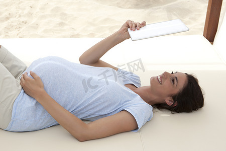 怀孕的中年妇女躺在海滩的沙发上抬头看着数字平板电脑