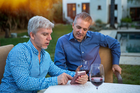 会议桌花摄影照片_两名成熟男子在花园聚会餐桌上使用智能手机