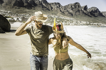 南非开普敦一对戴着草帽和羽毛头饰的中年夫妇在海滩上跑步