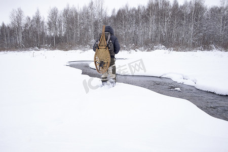 雪摄影照片_俄罗斯乌拉尔穿着传统雪鞋的中年男子在河流覆盖的雪地上的背影