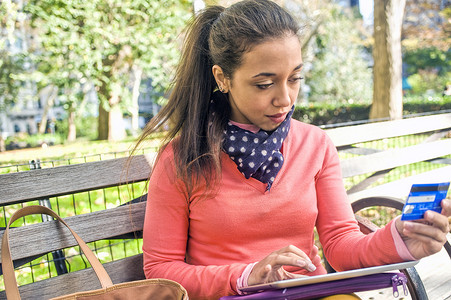 坐在公园长椅上的年轻女子使用带有数字平板电脑的信用卡
