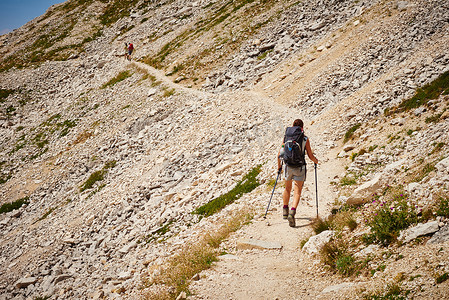 行走的力量摄影照片_徒步旅行者背着拐杖行走在山路上的后景奥地利