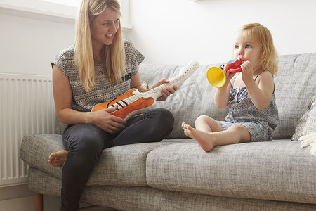 蹒跚学步的女孩子和妈妈在沙发上玩玩具小号和吉他