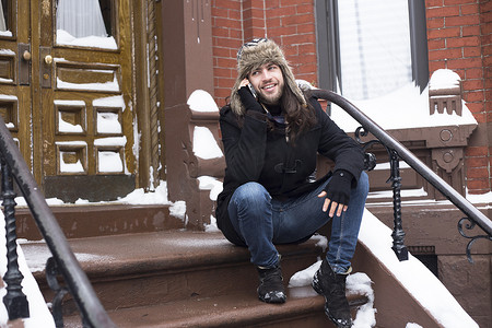智能造摄影照片_一名年轻人在积雪覆盖的前门台阶上用智能手机聊天