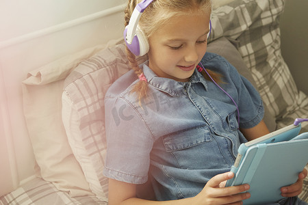 女孩戴着耳机坐在床上低头看着数字平板电脑微笑的截图