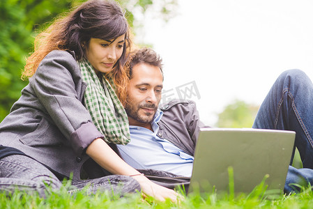 一对年轻夫妇坐在草地上用笔记本电脑俯视着