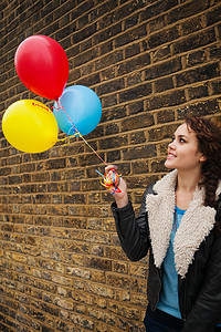 游戏红砖墙摄影照片_年轻女子拿着五颜六色的气球靠在砖墙上