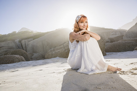 角色坐摄影照片_一名穿着白色连衣裙的女子坐在海滩上看着别处
