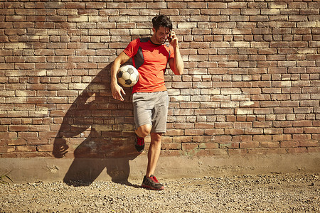 游戏红砖墙摄影照片_年轻的男子足球运动员在荒原上用智能手机聊天