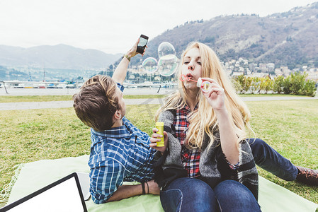 漂浮的泡泡摄影照片_意大利科莫湖年轻夫妇在野餐毯子上吹泡泡和拍摄泡泡