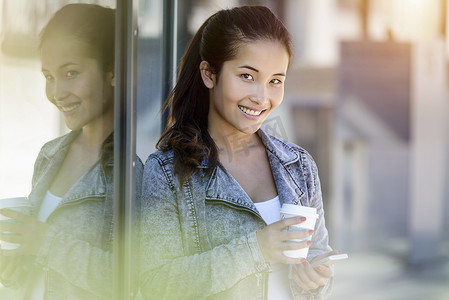 年轻女子的肖像手里拿着外卖咖啡和智能手机靠在公园大楼旁边