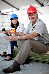建筑蓝红摄影照片_微笑的男女建筑工人坐下来手里拿着咖啡和蓝图