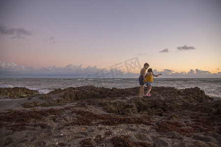 石凯摄影照片_女孩和兄弟在日出时跨过海滩岩石吹石保护区美国佛罗里达州朱庇特岛